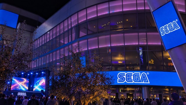 The front of the theater at night with SEGA on the band above the doors and the mast screen, and a play stage in front of the trees
